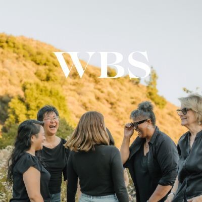 Group of women laughing and talking outdoors with hills in the background, wearing casual black outfits.
