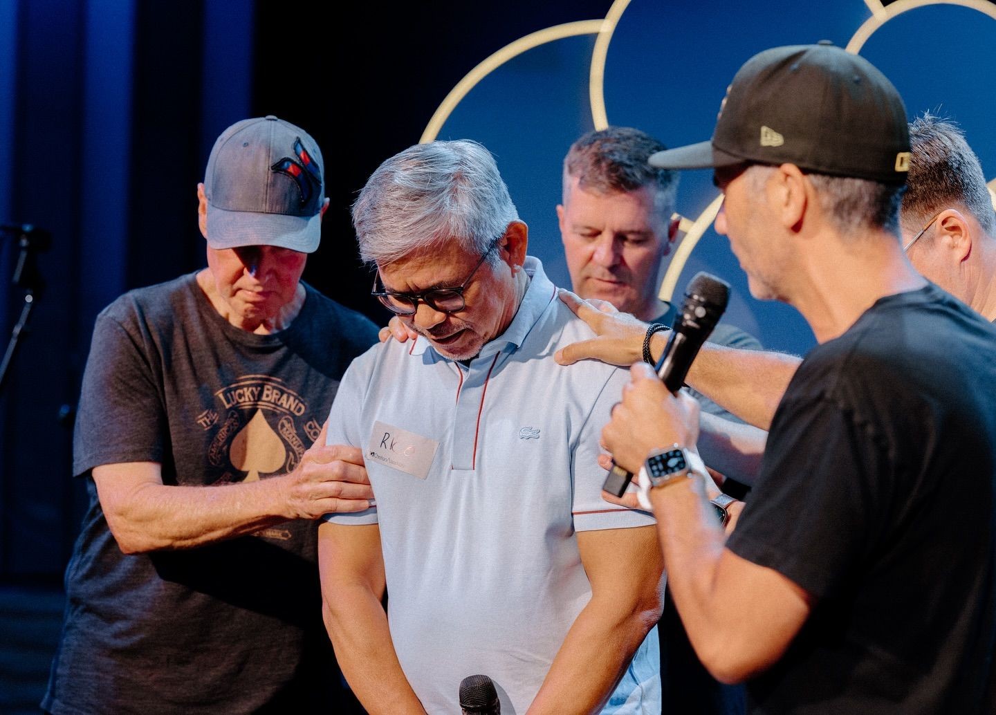 Group of men gathered in prayer, one holding a microphone, in a dimly lit room.