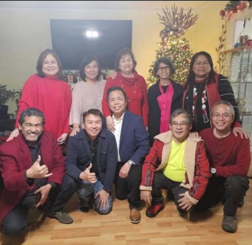 Group of people posing indoors with a Christmas tree in the background, some wearing red outfits.