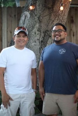 Two men smiling and standing outdoors in front of a tree, wearing casual T-shirts and shorts.