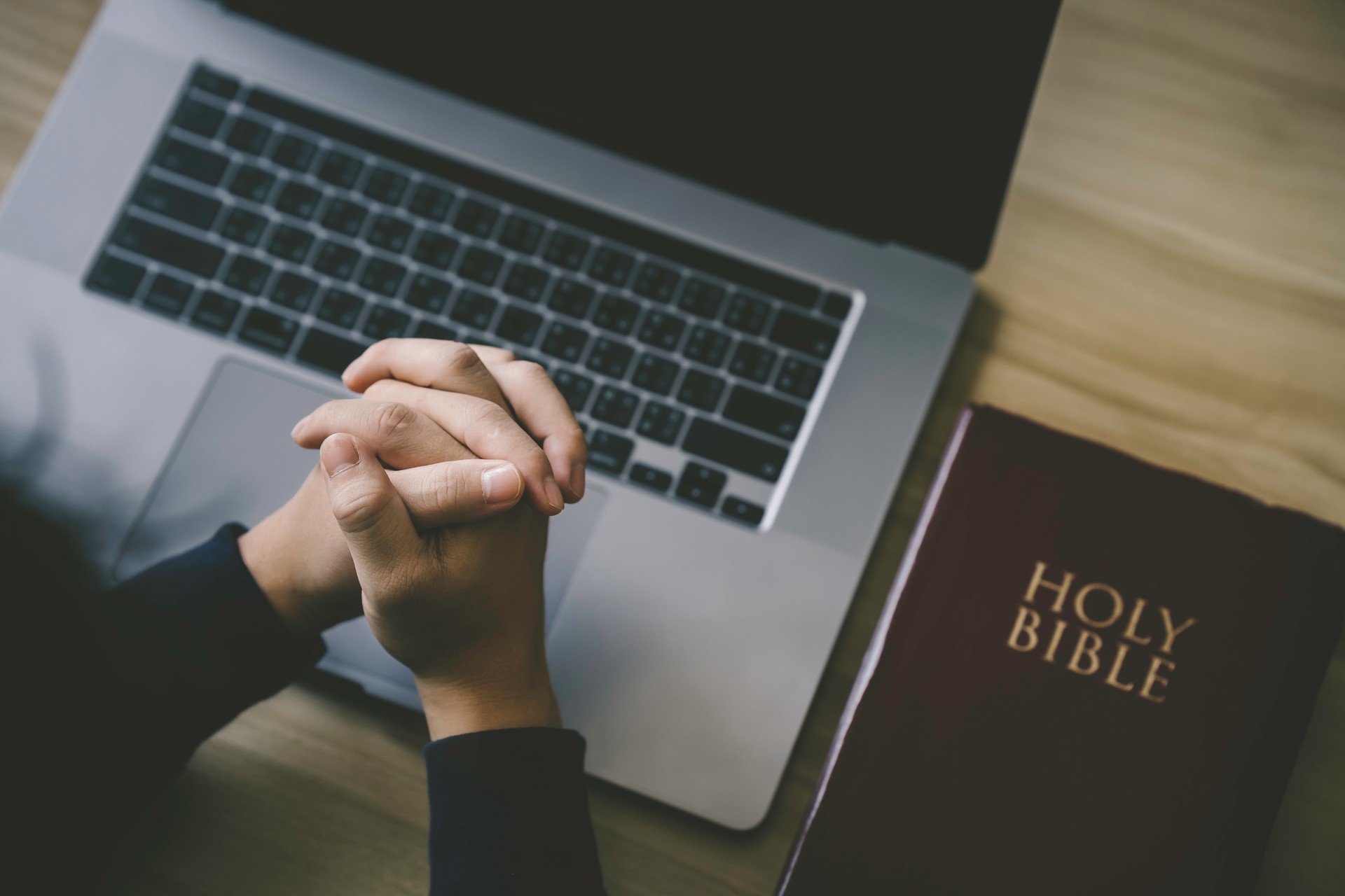Christian hands folded for prayer with a holy bible and laptop on a wooden table in church for worship online to god in the morning. person pray with faith, Spirituality, and religion concept